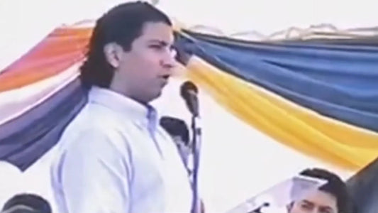 Man speaking at microphone outdoors with yellow, blue, and orange fabric backdrop, MEGA USA